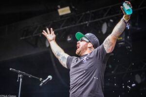 A tattooed man on stage with arms raised, wearing a black cap and sunglasses, holding a blue water bottle against a dark background.