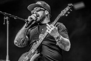 Male musician singing into a microphone while playing an electric guitar on stage; wearing a cap, glasses, and tattoos visible on arms (black-and-white photo).