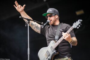Male guitarist on stage, wearing a black t‑shirt, cap, and sunglasses, raising his left hand while singing into a mic in front of him in a live show.