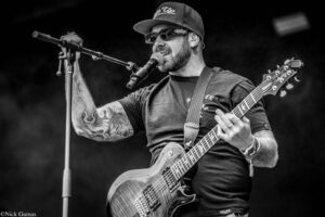 Guitarist wearing a cap and sunglasses sings into a microphone while playing an electric guitar on stage (black-and-white).