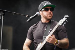 Musician performing on stage, wearing a black cap, sunglasses, and dark T-shirt while playing an electric guitar at a microphone stand.