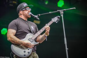 Guitarist wearing a black cap and sunglasses sings into a microphone while playing a silver electric guitar on stage.