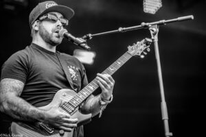 Male guitarist in a baseball cap and sunglasses sings into a microphone while playing a solid-body electric guitar on stage; visible tattoos on his arms.