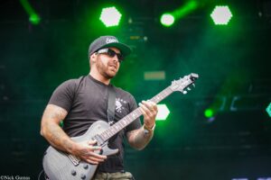 Guitarist wearing a black cap and sunglasses playing a gray electric guitar on a stage with bright green lights behind him.