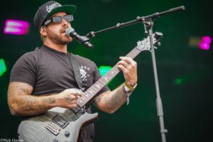 Male guitarist wearing a black cap and sunglasses performs on stage, singing into a microphone.