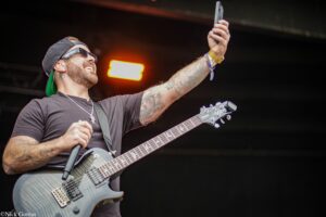 Guitarist on stage wearing a backward cap and sunglasses, raising a phone while playing a gray electric guitar; tattoos cover his arms.