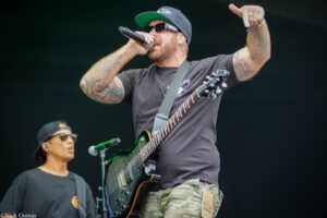 Male vocalist with a black guitar performs on stage, tattooed arms, dark cap and sunglasses, raising one arm mid-song at an outdoor concert.