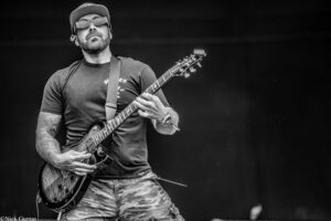 Male guitarist wearing a cap and sunglasses, playing an electric guitar on stage.