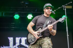 Male guitarist performing on stage, wearing a black t‑shirt, camouflage shorts, cap and sunglasses, playing an electric guitar with a microphone in front.