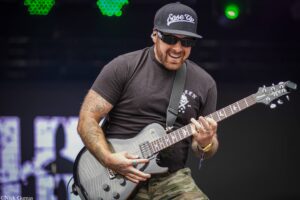 Guitarist performing on stage wearing a black cap and sunglasses, playing a gray electric guitar with a camouflage-pants musician look, smiling.