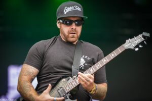 Male guitarist wearing a black cap and sunglasses, playing a dark electric guitar on stage with visible arm tattoos.