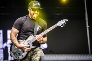 Guitarist wearing a black cap and sunglasses, playing a gray electric guitar on stage outdoors/at a concert, microphone cable visible.