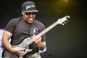 Guitarist wearing a black cap with a Goose Up logo, sunglasses, and tattoos, playing a gray electric guitar on stage.