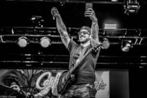 Rock guitarist on stage raises both arms in celebration, holding a microphone in one hand and a phone in the other, wearing sunglasses and camouflage pants (black and white).