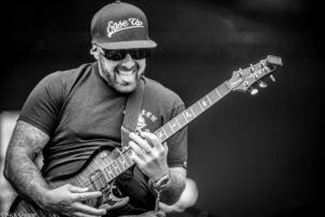 Guitarist wearing a cap and sunglasses, smiling as he plays an electric guitar on stage