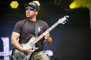 Guitarist wearing a black t-shirt, camo shorts, sunglasses, and a cap playing a gray electric guitar on stage.