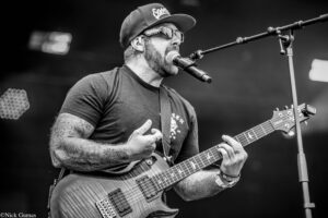 Black-and-white photo of a tattooed guitarist singing into a microphone while playing an electric guitar on stage at a concert.