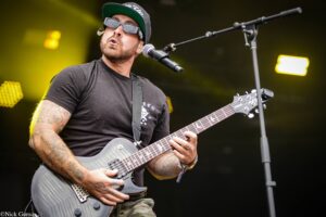 Male guitarist performing on stage with a gray electric guitar, wearing sunglasses and a cap, singing into a microphone.