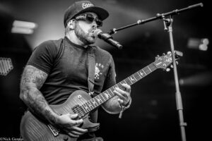 Male guitarist wearing a cap and sunglasses sings into a microphone while playing an electric guitar on stage (black and white).