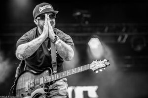 Black-and-white photo of a guitarist on stage with hands pressed together in a prayer-like pose, wearing a cap and sunglasses, visible tattoos on his arms.