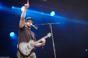 Guitarist on stage raising one index finger, wearing a cap, sunglasses, and tattoos, singing into a microphone under blue stage lights.
