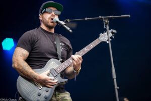Bearded male guitarist wearing a black T-shirt, sunglasses and a cap sings into a microphone while playing a gray electric guitar on stage.