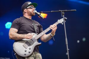 Guitarist performing on stage, wearing a black cap and sunglasses, singing into a microphone while playing a white electric guitar.
