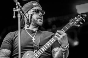 Black-and-white close-up of a guitarist wearing a backward cap and sunglasses, playing an electric guitar on stage.