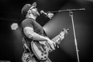 Black-and-white photo of a guitarist on stage, wearing a cap and sunglasses, singing into a microphone while playing an electric guitar.