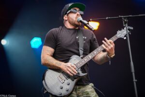 Male guitarist wearing a black cap and sunglasses, playing a gray electric guitar on stage with colorful lights in the background.