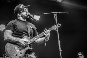 Black-and-white photo of a tattooed guitarist singing into a microphone while playing an electric guitar on stage, live performance context included.