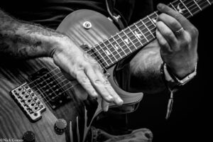 Close-up of a guitarist's hands on an electric guitar, fingers pressing the fretboard and plucking strings in black-and-white.