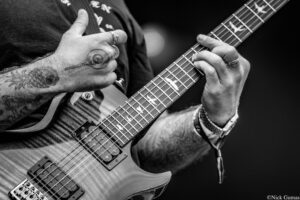 Close-up of a guitarist's tattooed hands playing an electric guitar, fingers pressing frets on a striped neck.