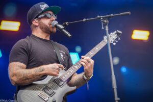 Male guitarist with tattoos playing a gray electric guitar on stage, singing into a microphone, wearing a black cap and sunglasses under blue stage lights.
