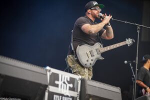 Male guitarist performing live on stage, wearing a black t-shirt and camouflage pants, tattooed arms, singing into a microphone while playing a gray electric guitar.