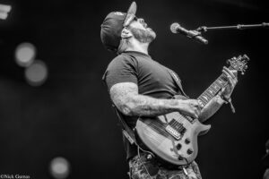 Male guitarist in a cap performing on stage, shouting into a microphone while playing an electric guitar; visible tattoos on his arms.