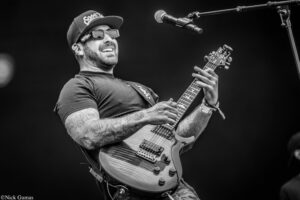 Male guitarist performing on stage, wearing a cap and sunglasses, playing an electric guitar with tattooed arms visible under a black-and-white photo