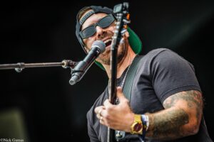 Male guitarist singing into a microphone on stage, wearing sunglasses and a backwards cap with a tattooed arm visible.