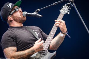 Male guitarist wearing a black cap and sunglasses, singing into a mic while playing a gray electric guitar on stage.