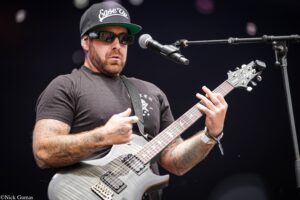 Male guitarist performing on stage, wearing a black cap, sunglasses, and a gray electric guitar with tattoos visible on arms.