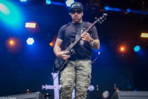 Guitarist in camo pants and black shirt playing an electric guitar on a neon-lit stage.