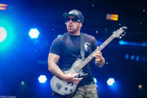 Guitarist on stage playing a gray electric guitar, wearing a black cap, sunglasses, and a black T-shirt; blue stage lights.