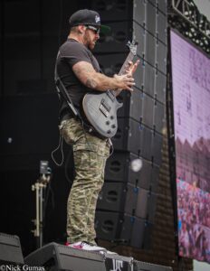 Tattooed guitarist in a black cap and sunglasses playing a gray electric guitar on a concert stage with large speaker stacks behind him.