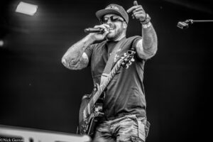 Black-and-white image of a male guitarist on stage, wearing a cap and sunglasses, singing into a handheld mic with tattoos visible on his arms and a guitar strapped across his chest.
