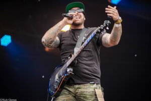 Male guitarist on stage wearing a green cap and sunglasses, singing into a microphone while playing a blue electric guitar.