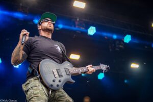 Guitarist on stage wearing a black shirt, camo shorts, a green cap, sunglasses, playing a gray electric guitar while holding a mic.