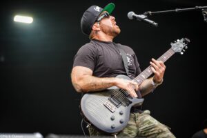 Guitarist performing on stage, wearing a black cap, sunglasses, and camouflage pants, playing a grey electric guitar into a microphone.