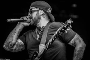 Male guitarist and vocalist performing on stage, wearing a backward cap, sunglasses, and visible tattoos on his arms.