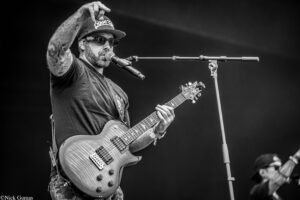 Male guitarist performing on stage, wearing sunglasses and a cap, singing into a microphone while strumming a sunburst electric guitar; visible tattoos on his arm and a dynamic, focused pose.