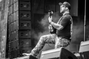 Guitarist performing on stage, wearing a cap and sunglasses, mid-kick with one leg on a stage monitor while playing a guitar (black and white).